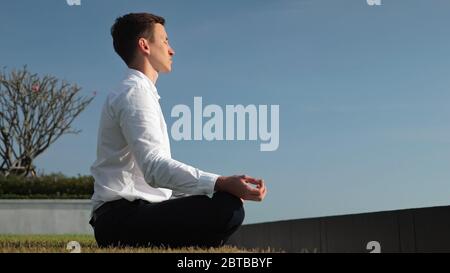 young businessman in white shirt meditates sitting in lotus pose on hotel terrace at sunlight against blue sky side view Stock Photo