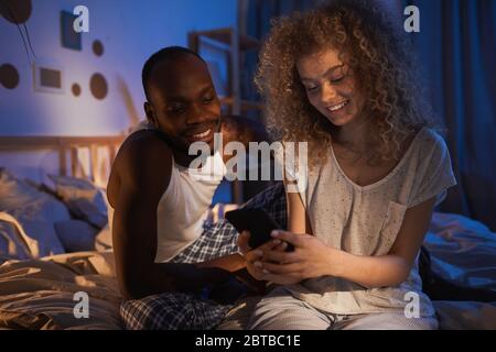 Portrait of young mixed-race couple sitting on bed at night and smiling happily while looking at smartphone screen Stock Photo