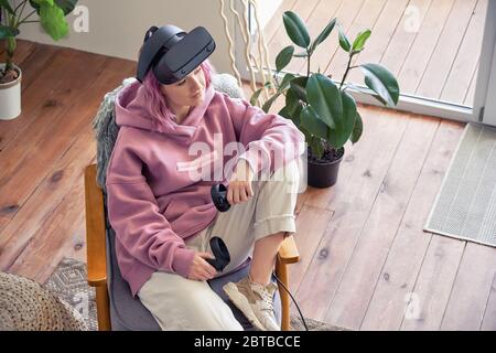 Fashion teen girl wear vr headset hold controllers sit relax in chair, top view. Stock Photo