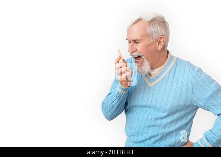 Freestyle. Senior bearded man in sweater standing isolated on white shouting at smartphone smiling happy side view Stock Photo