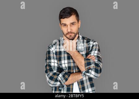 Freestyle. Young man in checkered shirt standing isolated on grey crossed arms touching chin pensive Stock Photo