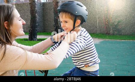 Portrait of smiling young mother adjusting and fastening her son protective helmet buckles before riding bicycle Stock Photo