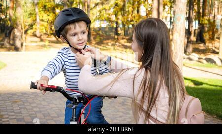 Portrait of beautiful young mother adjusting safety helmet on her son's head before riding a bicycle at park Stock Photo