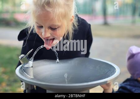 Cute adorable caucasian blond little thirsty school girl drinking water from public potable fountain faucet in city park on bright hot summer day