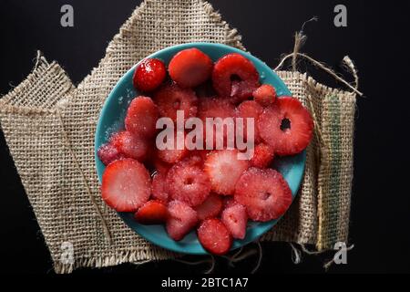 Chopped strawberries with sugar on a blue plate isolated on a black desk and retro canvas sack. Summer and season food concept Stock Photo