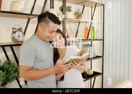 Cheerful smiling young couple standing at shelf and reading book together Stock Photo