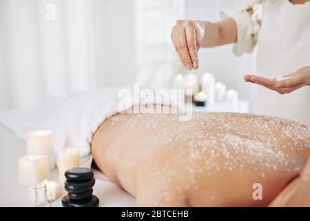 Beautician applying salt scrub on back of male client to get rid of dead skin before massage Stock Photo