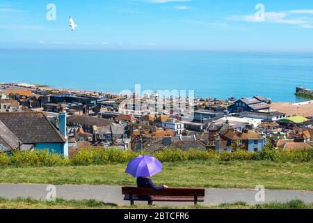 Hastings, a girl with a purple parasol sitting in the sun on West Hill overlooking the view to the Old Town Harbour and sea, Sussex, UK 20th May 2020 Stock Photo