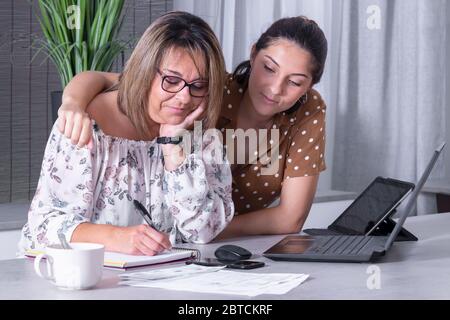 Smiling mature woman writing on a notebook surrounded by a laptop and a tablet while being embraced by her daughter. Home office and family concept. Stock Photo
