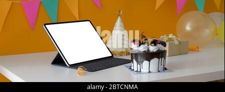 Online birthday party concept with blank screen tablet, cake, party hat and present box on white table with decorations on yellow wall Stock Photo