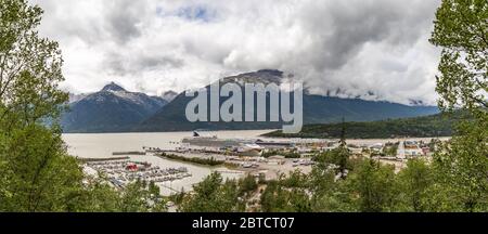 Panoramic view of Skagway port with Norwegian Jewel docked there. Mountains and clouds in the background. Forest in the foreground. Stock Photo