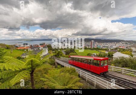 Red cable car moving up the mountain in the foreground and panorama of Wellington in the background. Beautiful blue sky covered with clouds. Stock Photo