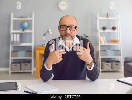 Mature man with a gray beard in spectacled looks at the camera, speaks with his partner sitting at a table in the office at home. An old male teacher Stock Photo