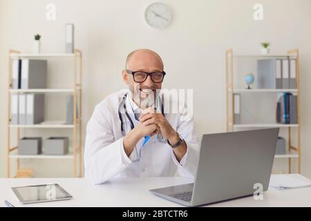 Mature doctor.A middle-aged confident doctor smiles while sitting at a table with a laptop at the workplace in the clinic office. Stock Photo