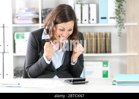 Excited adult executive woman celebrating checking result on calculator sitting on a desk at office Stock Photo