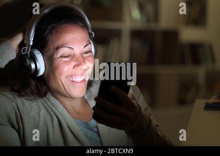 Happy adult woman with headphones listens music on smart phone with lighted screen in the livingroom at night at home Stock Photo