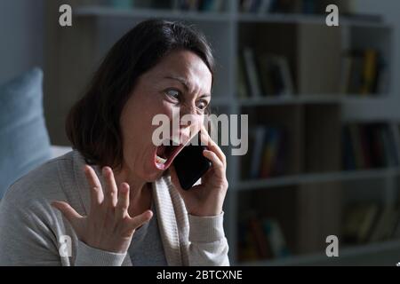 Angry adult woman screaming calling on smart phone sitting in the livingroom at night at home Stock Photo