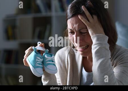 Sad mother crying missing her daughter after miscarriage holding new baby shoes at night at home Stock Photo