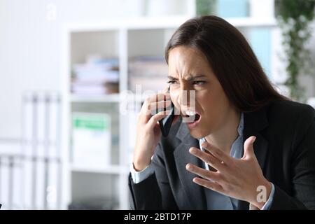 Angry executive woman arguing calling on smart phone sitting on a desk at the office Stock Photo
