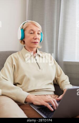 Serious senior woman in headphones sitting on sofa and working online using her laptop computer Stock Photo