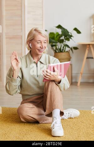 Mature woman smiling and gesturing while sitting on the floor and using tablet pc for online conversation Stock Photo
