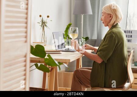 Side view of mature woman sitting at the table with wineglass of wine and using laptop at home Stock Photo