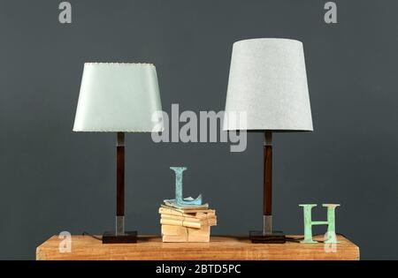 Two vintage table lamps with off white cylindrical shades on an old desk with books and metal letters against a grey wall Stock Photo