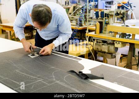Tailor bending over a workbench cutting fabric on which a pattern has been stencilled in a large spacious industrial workshop Stock Photo