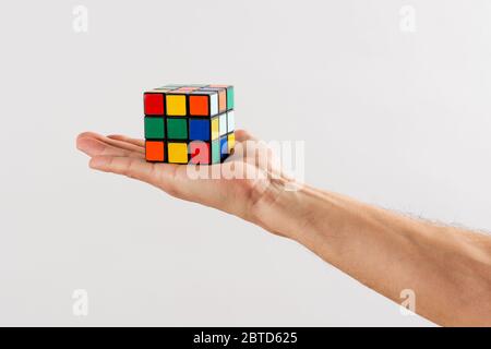 Man holding up an unsolved Rubiks Cube puzzle on the palm of his hand extended over a white wall background Stock Photo