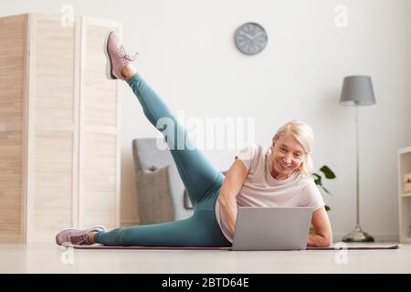 Mature woman lying on the floor on exercise mat and stretching her leg while looking sports training on laptop at home Stock Photo