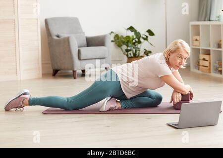 Senior woman doing exercises on exercise mat and watching these exercises on laptop at home Stock Photo