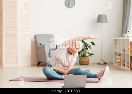 Senior woman sitting on the floor on exercise mat and stretching her hands while watching sports training on laptop at home Stock Photo