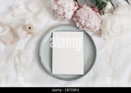 Wedding still life, menu mockup scene scene. Greeting card on grey porcelain plate. Silk ribbon and pink peony flowers on white linen table cloth. Vin Stock Photo