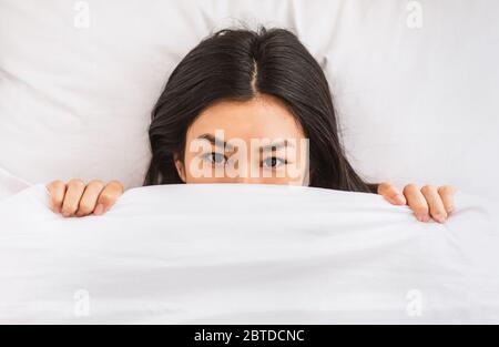 Asian Woman Peeking Out Of Blanket Waking Up In Bed Stock Photo