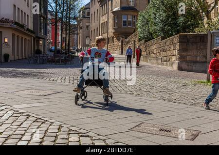 A boy steers his grandmother's rollator over the cobblestones of a lane near Burtscheider Markt in Aachen, an elderly woman using two crutches walking in the background. April 2011. Stock Photo