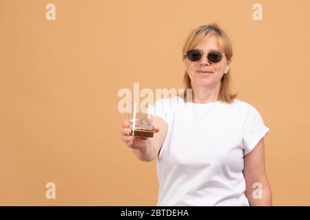 Happy smiling elderly woman in white t-shirt and round sunglasses holds glass with ice cube and looking at the camera, isolated on orange background Stock Photo