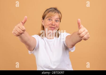 Smiling mature woman in t-shirt isolated on orange studio background show thumbs up recommend service Stock Photo