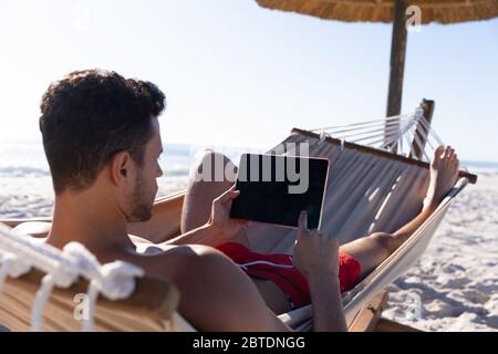 Caucasian man lying on a hammock and using a digital tablet at the beach. Stock Photo