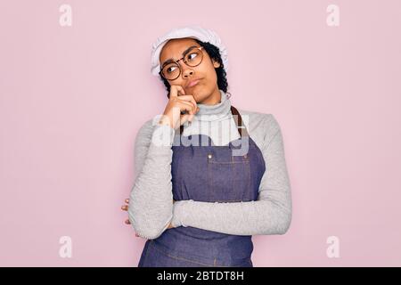 Young african american curly baker woman wearing apron and cap over pink background with hand on chin thinking about question, pensive expression. Smi Stock Photo