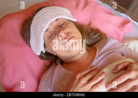 Middle aged woman has a fever or abnormal blood pressure. 40 years women lying in the bed with wet compress on a head. She is unwell and sick. Girl cl Stock Photo