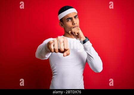 Young handsome african american sportsman wearing sportswear over red background Punching fist to fight, aggressive and angry attack, threat and viole Stock Photo