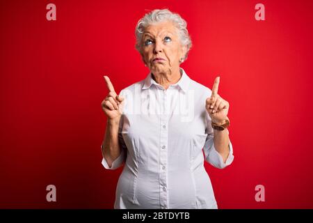 Senior beautiful woman wearing elegant shirt standing over isolated red background Pointing up looking sad and upset, indicating direction with finger Stock Photo