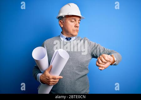 Middle age handsome grey-haired architect man wearing safety helmet holding blueprints Checking the time on wrist watch, relaxed and confident Stock Photo