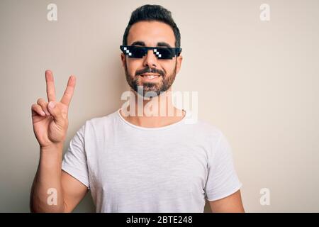Young handsome man with beard wearing funny thug life sunglasses over white background smiling with happy face winking at the camera doing victory sig Stock Photo