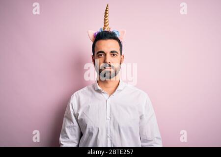 Young handsome man with beard wearing funny unicorn diadem over pink background with serious expression on face. Simple and natural looking at the cam Stock Photo