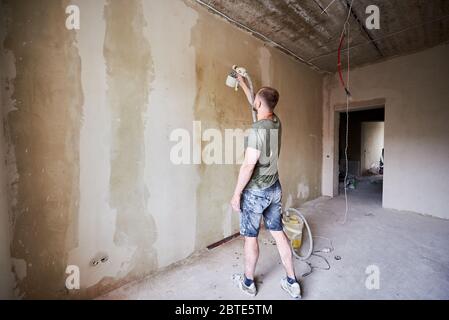 Back view of guy holding in hand tool spray of paint and painting a wall in the daytime. Male is dressed in paint-smeared clothing. House remodel. Stock Photo