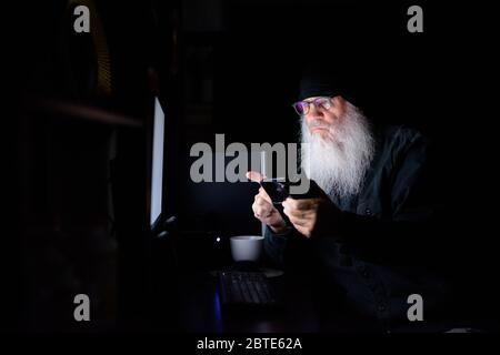 Mature bearded hipster man using phone while working overtime at home late at night in the dark Stock Photo