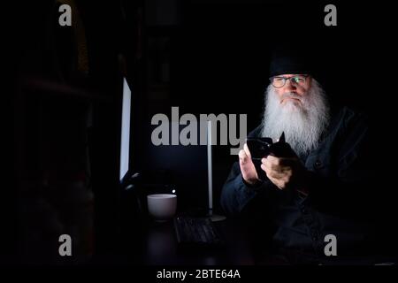 Mature bearded hipster man using phone while working overtime at home late at night in the dark Stock Photo