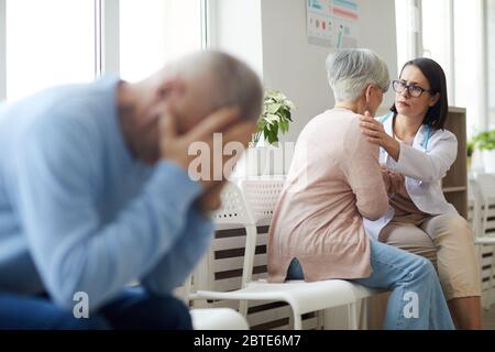 Back view portrait of senior woman talking to female doctor with blurred shape of frustrated husband in foreground, copy space Stock Photo