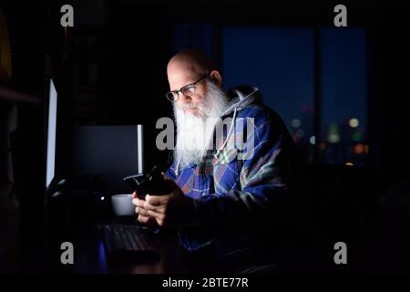 Mature bald bearded hipster man with eyeglasses using phone and working overtime at home late at night Stock Photo